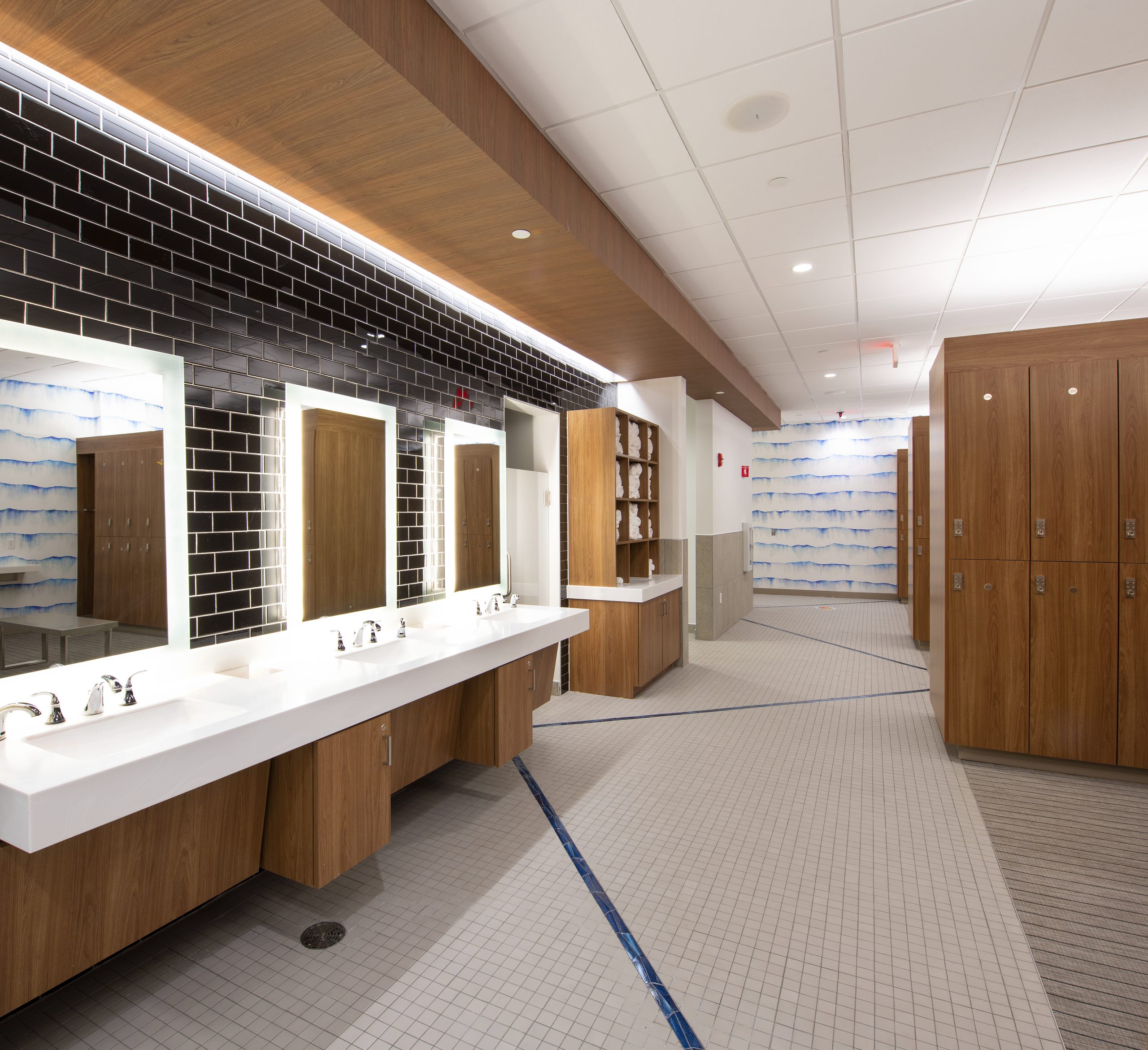 Luxury locker room vanity area with backlit mirrors, warm wood cabinetry, and white quartz countertops at Lake Nona Performance Club's steam and sauna locker rooms in Lake Nona, FL